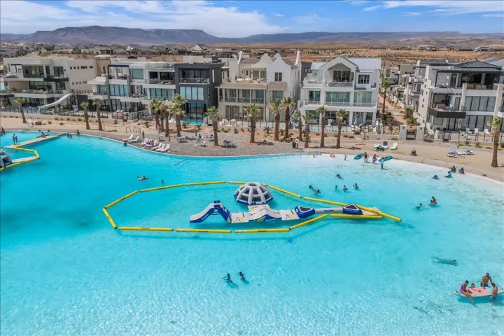 Expansive artificial lagoon with a sandy beach and inflatable water features, bordered by contemporary multi-story residences