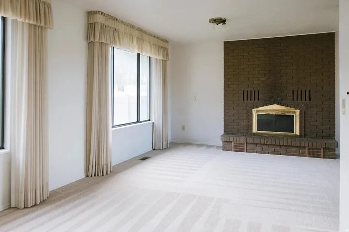 Brick fireplace with a brass firebox, light-toned wall-to-wall carpeting, and large windows with privacy treatments