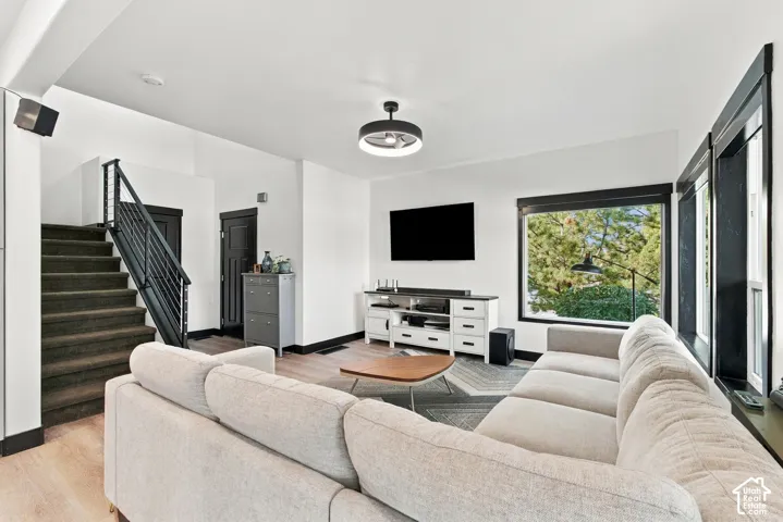Spacious living area featuring light wood-finish flooring, a contemporary ceiling fan with integrated lighting, and large windows with dark trim