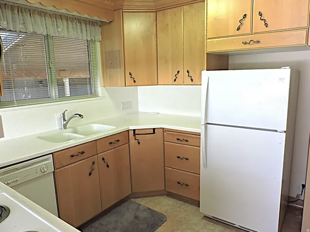 Kitchen featuring wood cabinetry, solid surface countertops, double basin sink with chrome faucet, and white appliances including a dishwasher and refrigerator