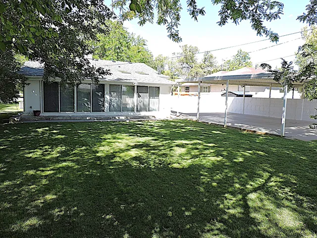 Expansive grassy yard with mature trees, a multi-panel sliding glass door entry, and a covered carport with concrete paving