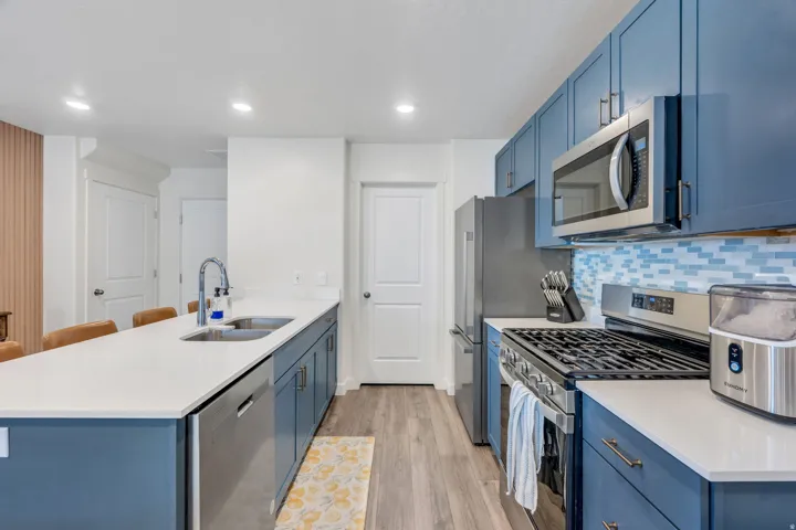 Stainless steel appliances, blue cabinetry, light-toned backsplash, light wood-finish flooring, and a kitchen island with sink