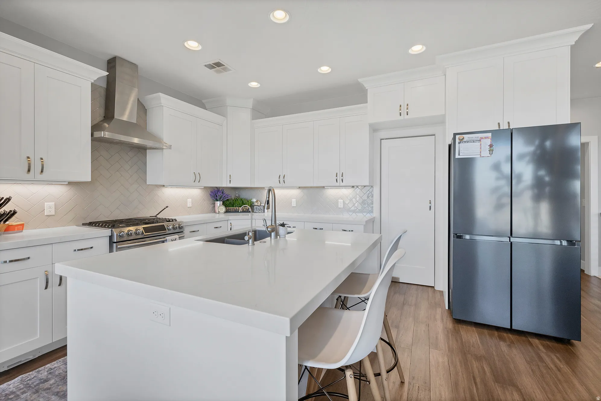 White cabinetry with crown molding, stainless steel range hood, herringbone tile backsplash, recessed lighting, and wood-finish flooring