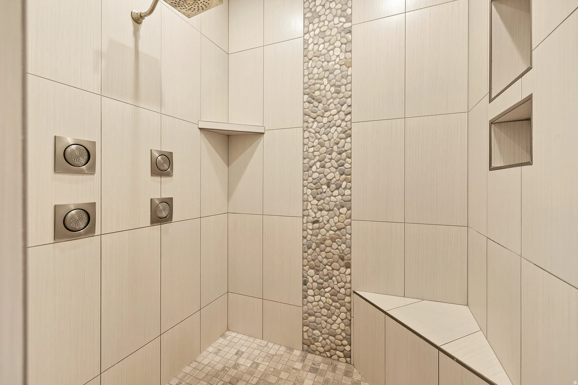Tiled shower with a pebble accent wall, built-in bench, recessed shelving, and multiple shower controls