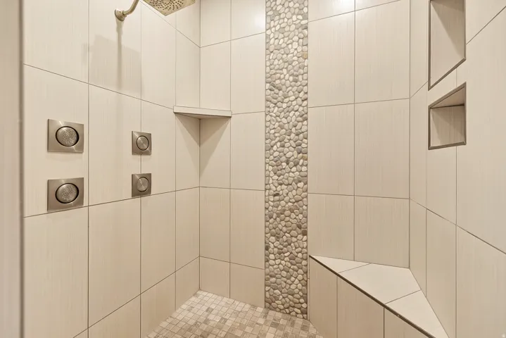 Tiled shower with a pebble accent wall, built-in bench, recessed shelving, and multiple shower controls