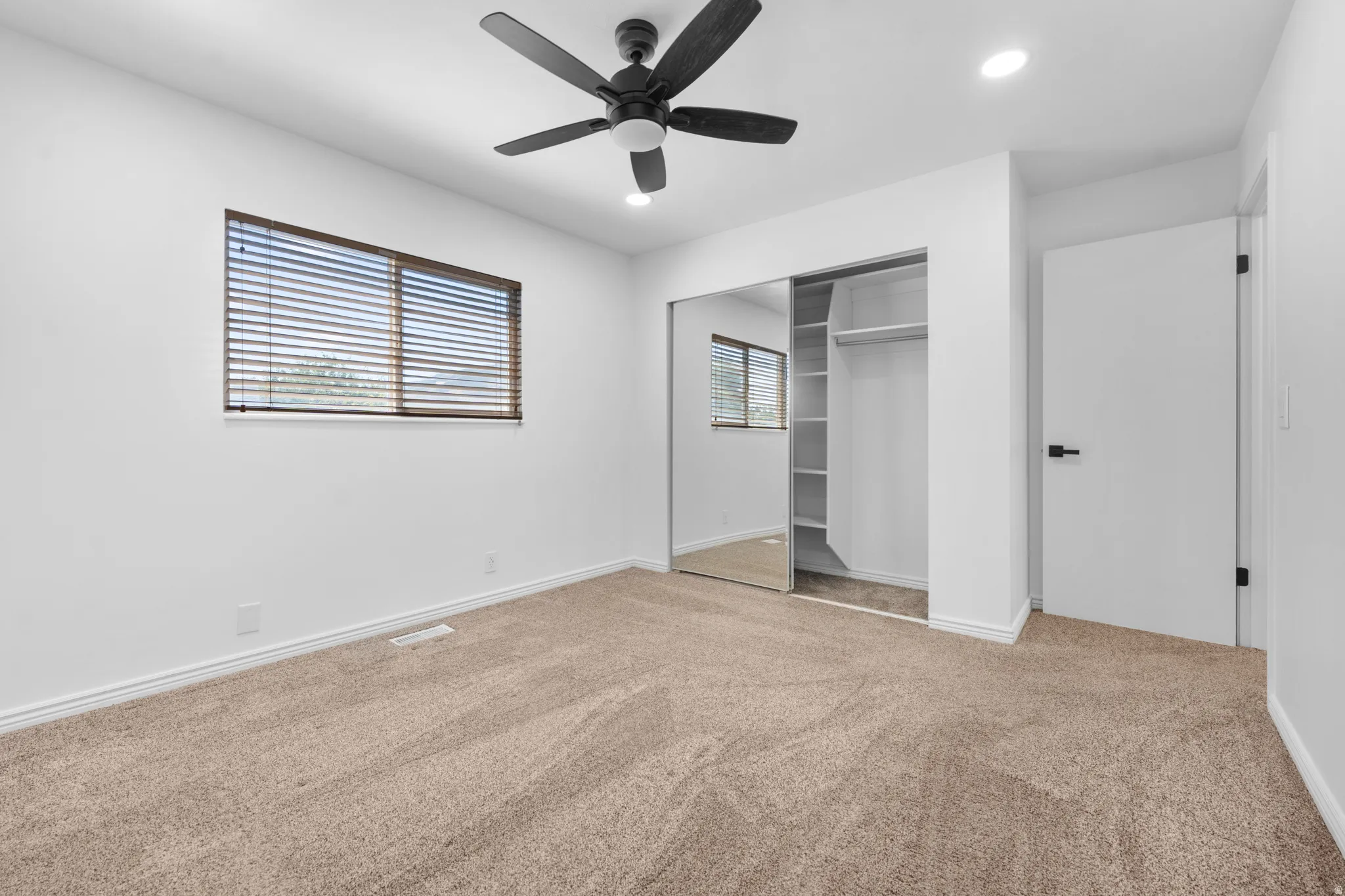 Spacious room featuring neutral carpeting, a black ceiling fan with integrated lighting, a window with horizontal blinds, and a built-in closet with mirrored sliding doors and shelving