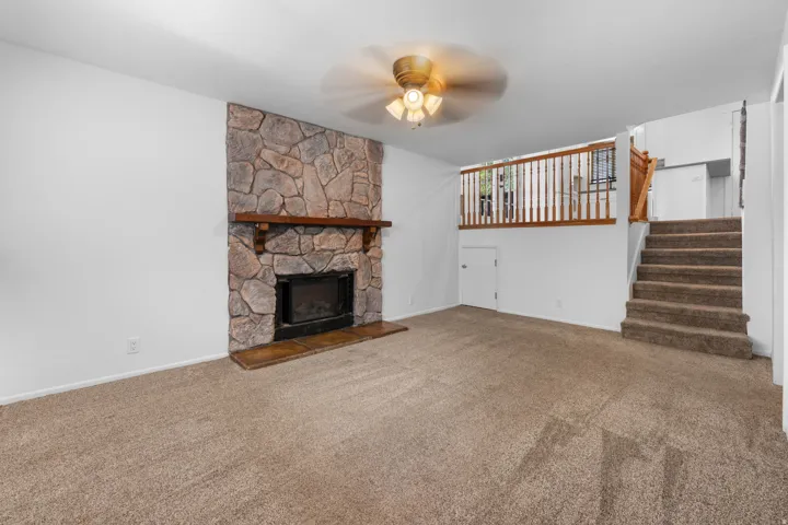 Stone-clad fireplace with a wood mantle, ceiling fan, carpeted flooring, and a built-in staircase with wood railing. Entrance to crawl space.
