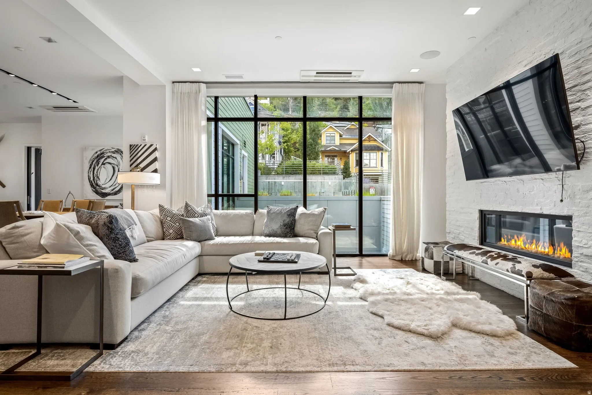 Expansive living area with wood-finish flooring, a full-height window, a linear fireplace, and a textured white brick accent wall