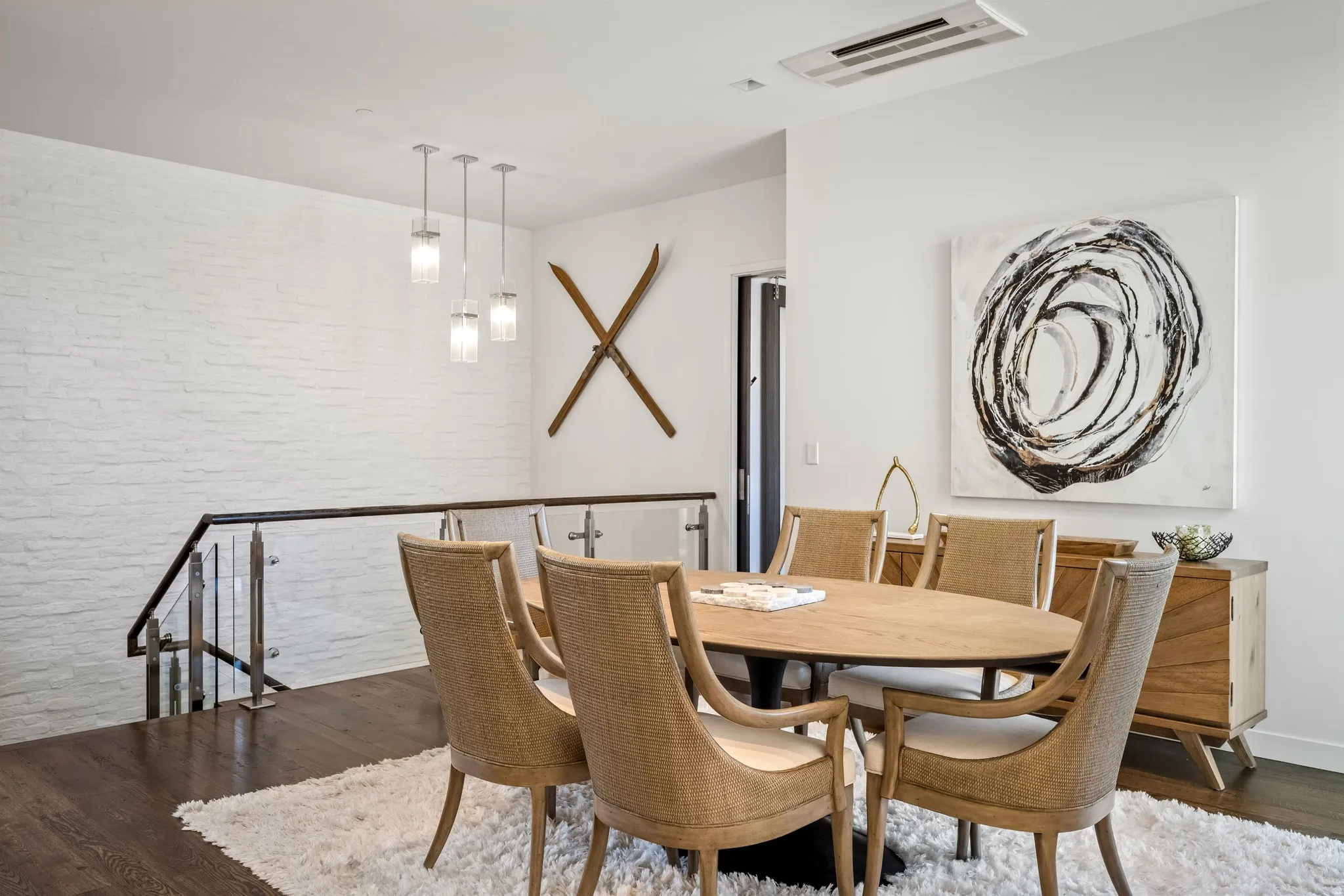 White painted brick accent wall, sleek glass and metal staircase railing, dark wood-finish flooring, contemporary pendant lighting, and a built-in wood credenza