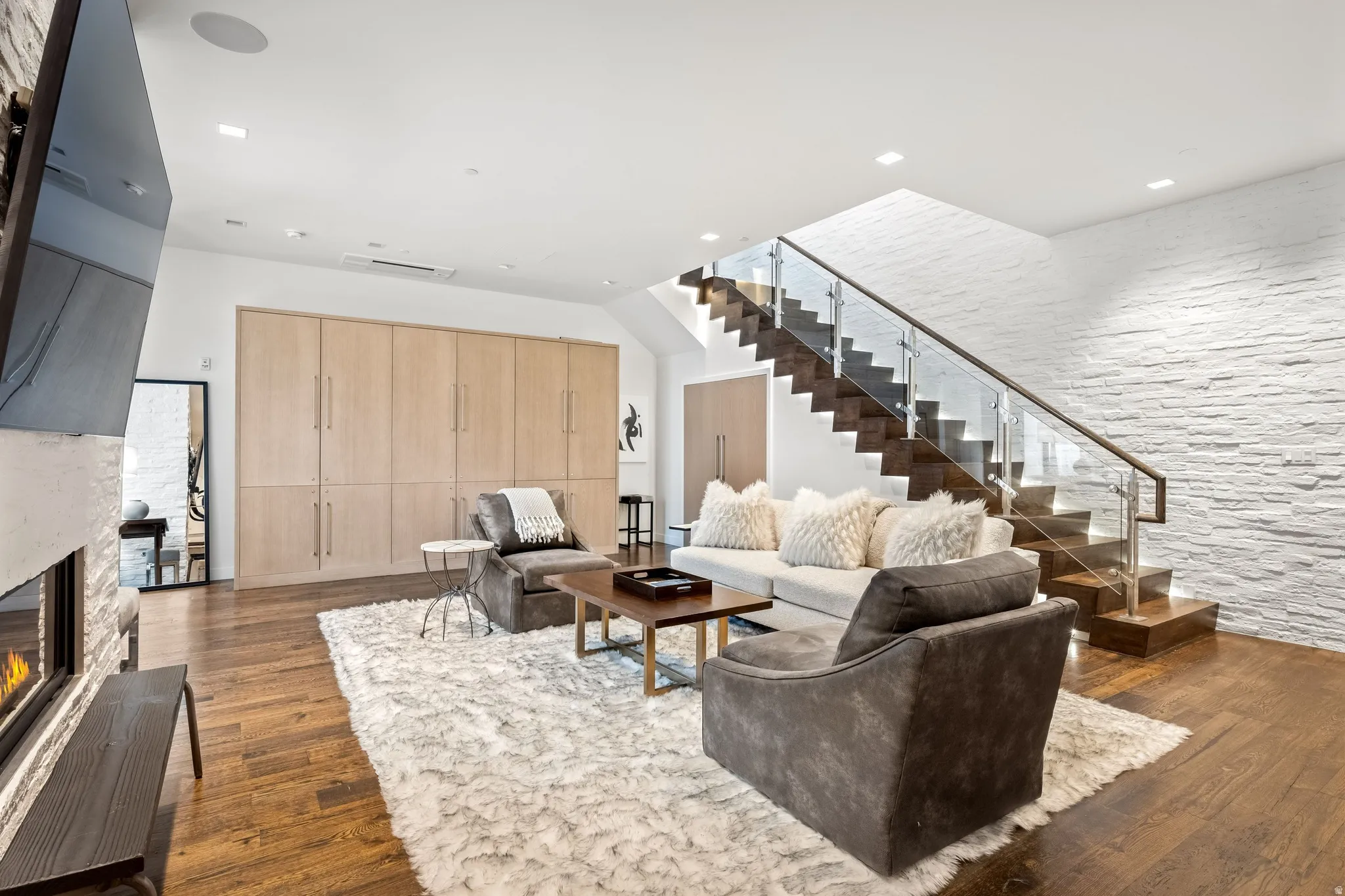 Hardwood flooring, linear gas fireplace with stone surround, wood-paneled built-in storage, open-tread staircase with glass railing, and a textured accent wall