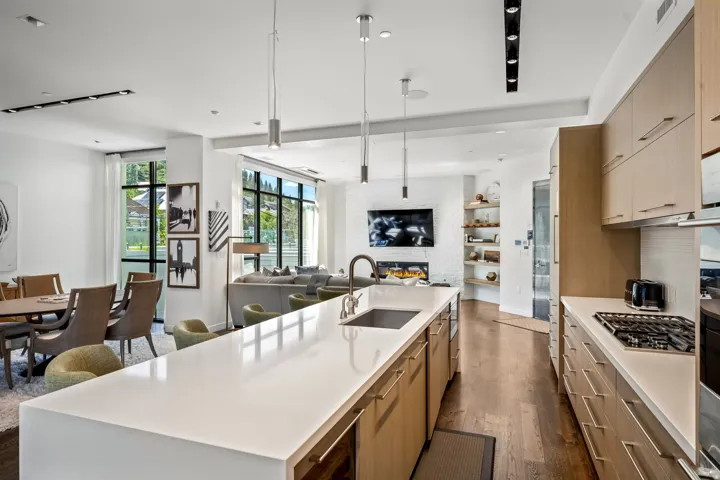 Open-concept living space with wood-finish flooring, a kitchen island with integrated sink, modern pendant lighting, extensive built-in cabinetry, and a gas cooktop