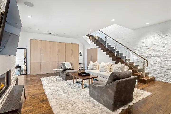 Hardwood flooring, linear gas fireplace with stone surround, wood-paneled built-in storage, open-tread staircase with glass railing, and a textured accent wall
