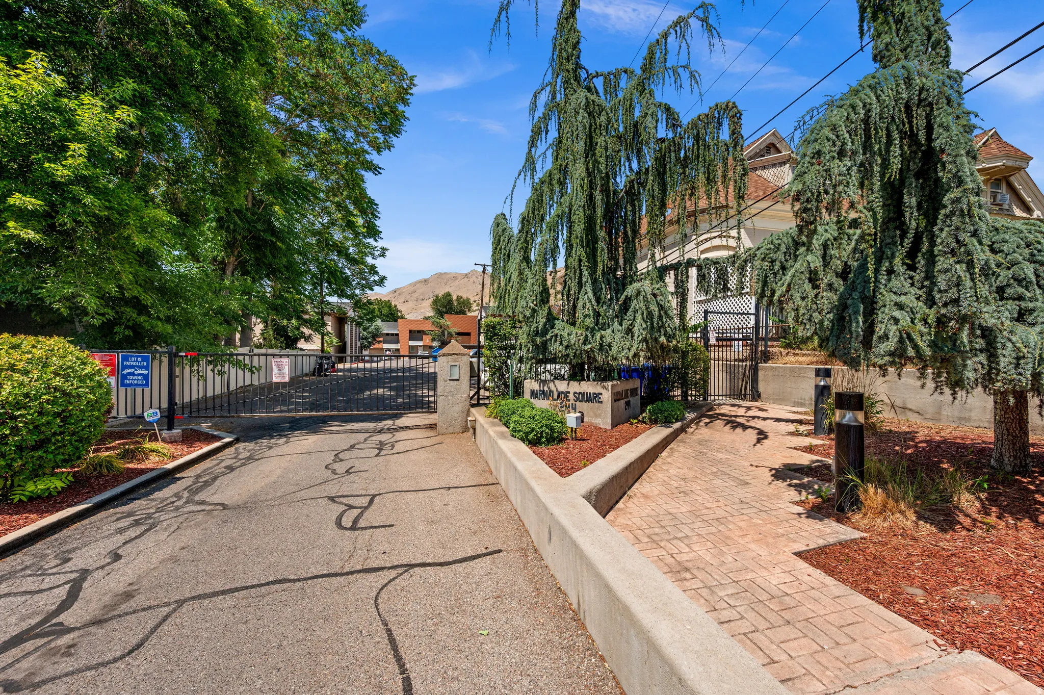 Gated property entrance with a brick paver walkway, mature landscaping, and an architectural building with multiple dormer windows