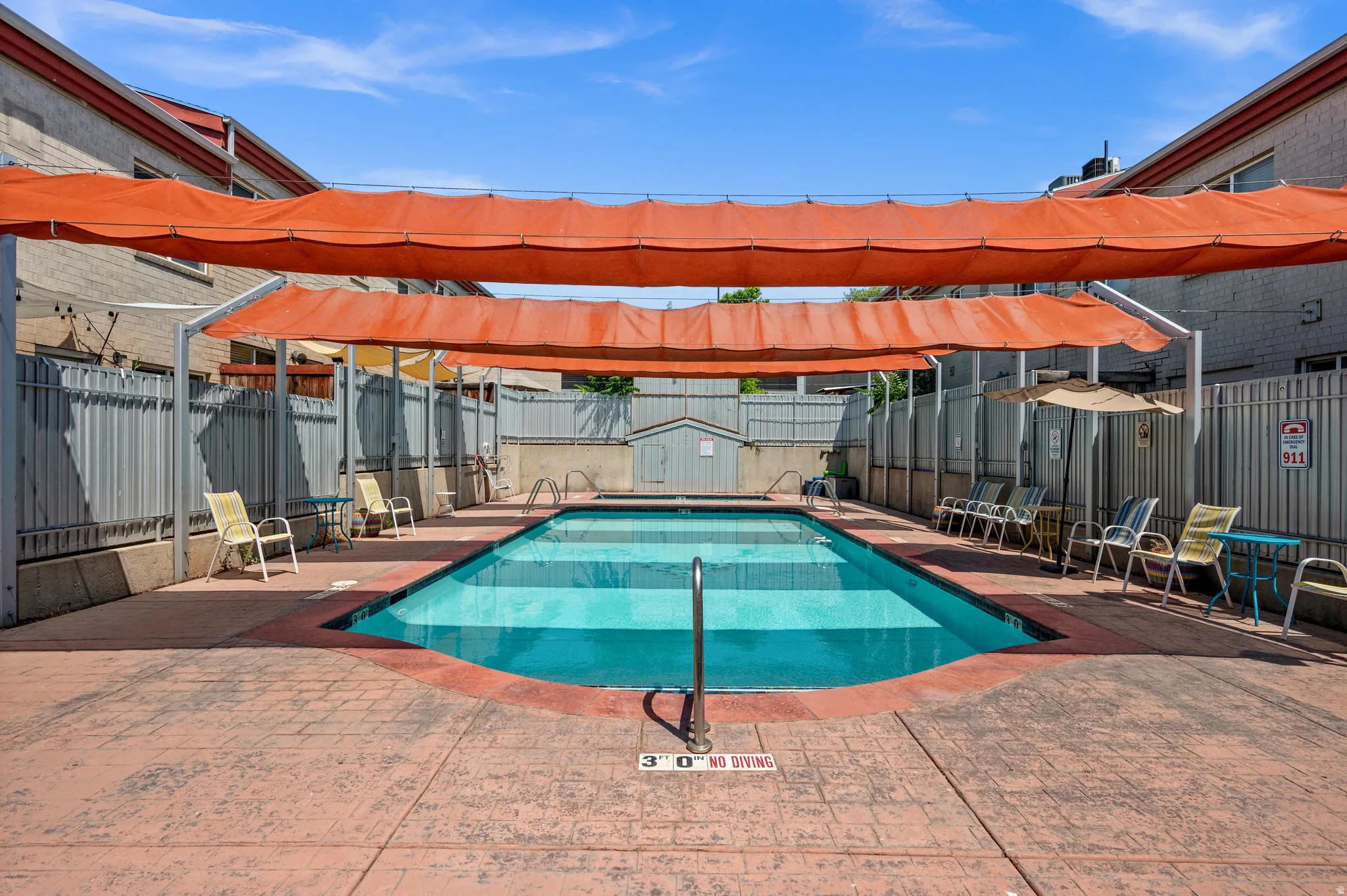 Outdoor swimming pool with a textured concrete deck, shaded by orange fabric canopies, and surrounded by fencing