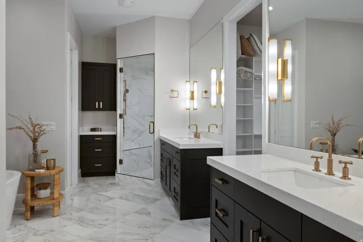 Dual dark wood vanities with white countertops, gold-toned fixtures, a walk-in shower with clear glass enclosure, and large-format floor tiling