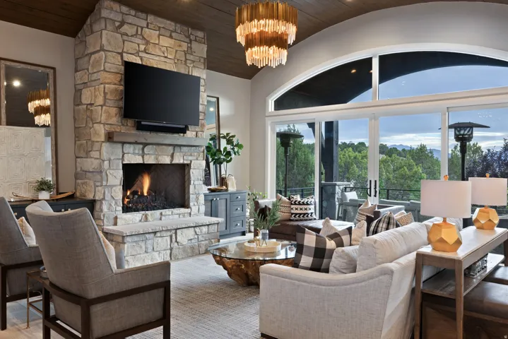 Sophisticated great room showcasing a two-sided fireplace with stone surround, wall-mounted flat-screen television, tiered crystal chandelier, and expansive arched windows framing natural landscape vistas