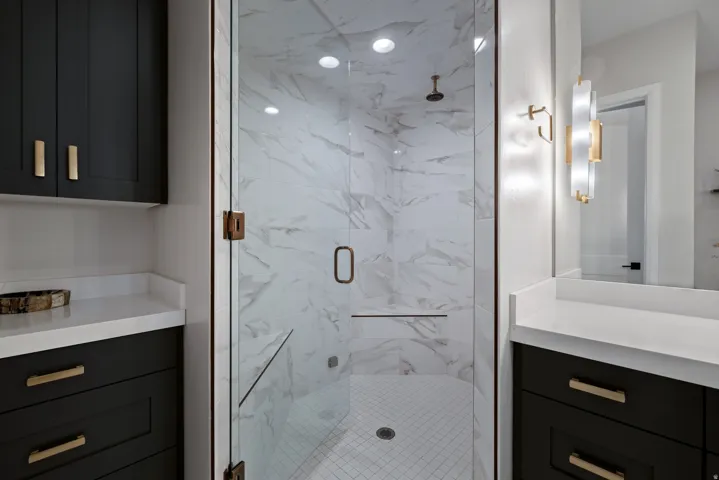 Glass-enclosed shower with white tiled walls and floor, complemented by dark cabinetry with gold-toned hardware, white countertops, and a contemporary wall sconce