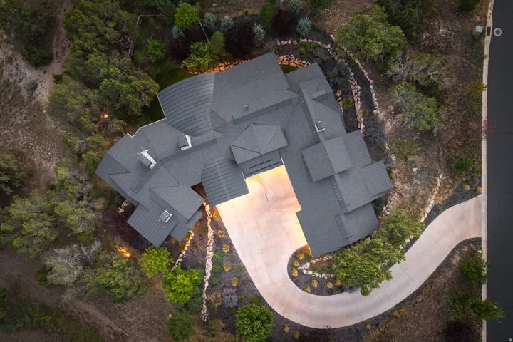 Multi-level roofline with dark shingles, curved concrete driveway, integrated exterior lighting, and natural rock landscaping