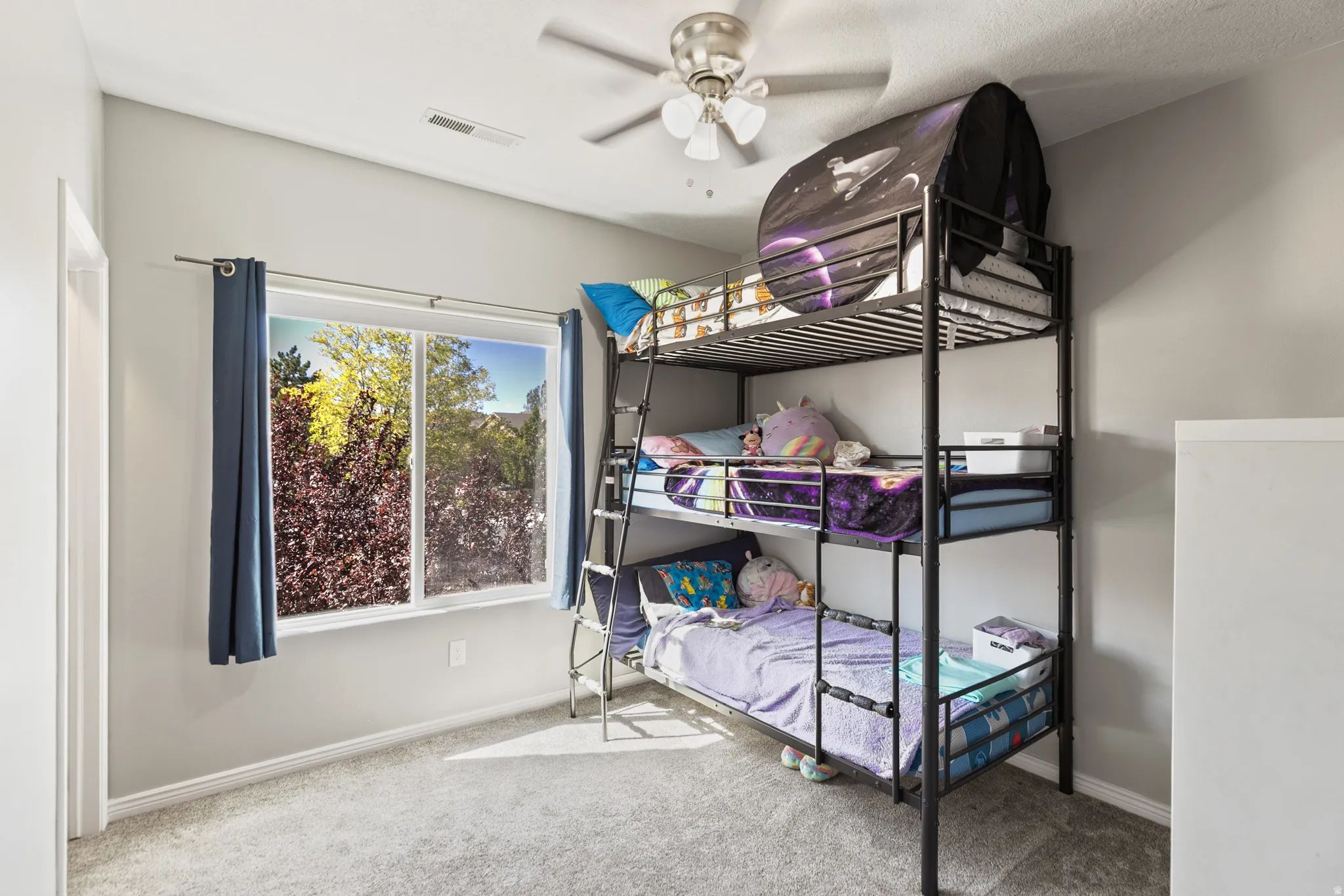 Room with light gray walls, a window featuring exterior landscaping, light gray carpet, and a ceiling fan with integrated lighting