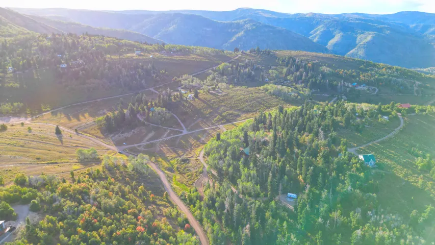 Expansive mountain landscape with diverse tree coverage, winding unpaved roads, and multiple distant structures