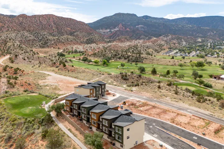 Contemporary townhomes with multi-story facades, dark-toned sloped roofs, and paved roadways, situated alongside a verdant golf course and rugged mountainous terrain