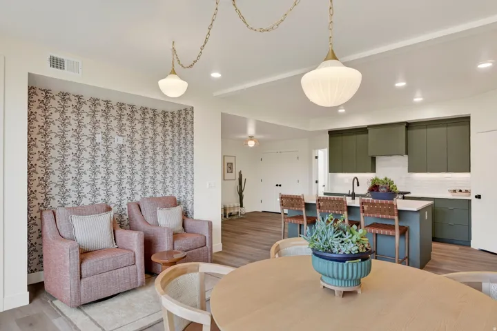 Recessed lighting and pendant fixtures illuminate the space, featuring wood-finish flooring, a kitchen island with seating, a white subway tile backsplash, and green cabinetry