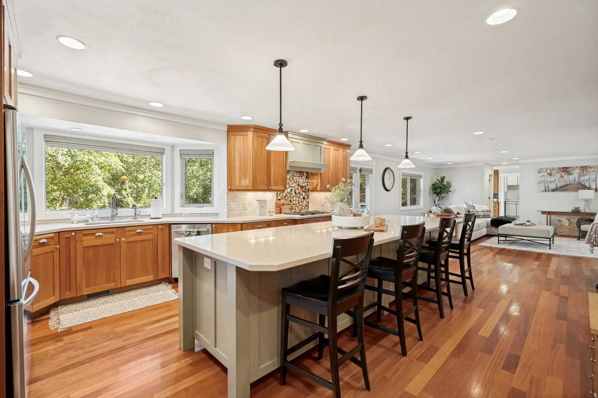 Gourmet kitchen featuring warm wood cabinetry, white quartz countertops, a large center island with seating, pendant lighting, stainless steel appliances, and hardwood flooring throughout the open-concept space with abundant natural light from multiple wi