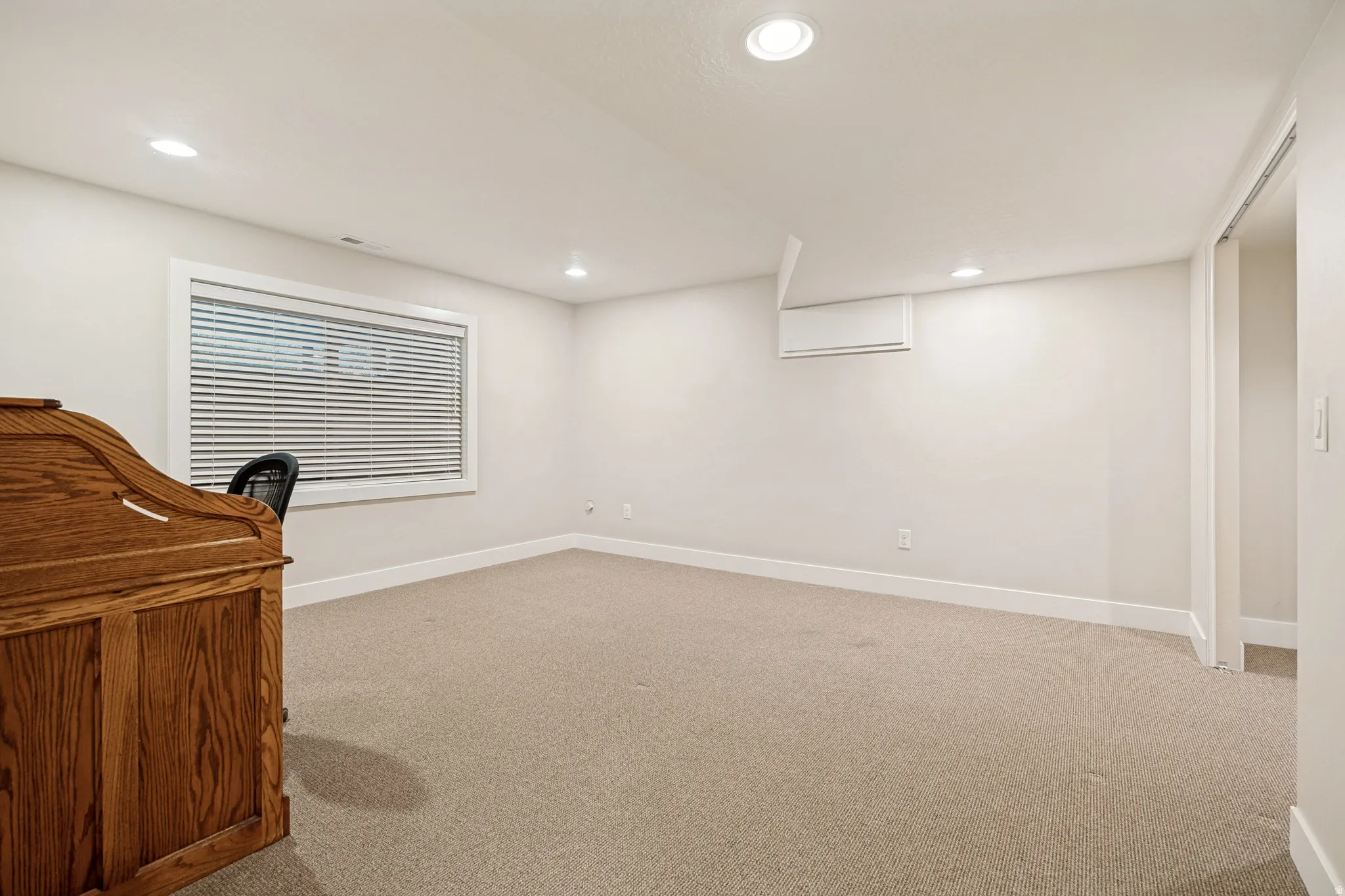 Recessed ceiling lighting, window with blinds, light-toned carpet flooring, white walls, and a wall-mounted mini-split unit