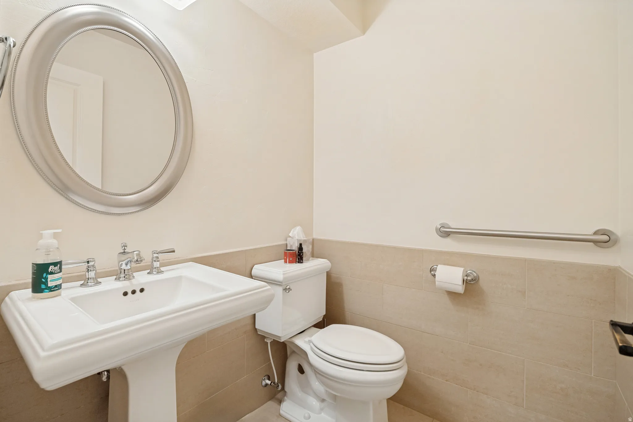 Pedestal sink with chrome faucet, oval framed mirror, wall-mounted grab bar, and tiled wainscoting