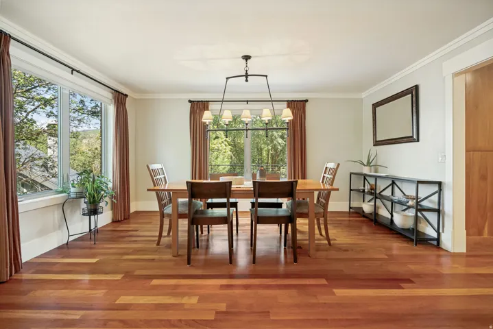 Elegant dining room showcasing rich wood-finish flooring, a large dining table with seating for six, and abundant natural light from multi-pane windows framed with warm drapery