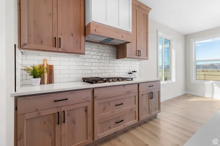Wood-finish cabinetry with matte black hardware, white subway tile backsplash, light-toned countertops, built-in range, and light wood-finish flooring