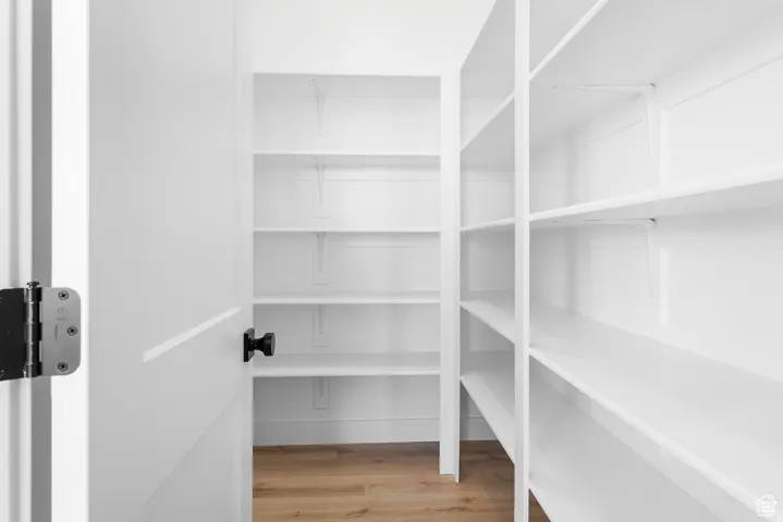 Walk-in pantry featuring white shelving, wood-finish flooring, and a black doorknob
