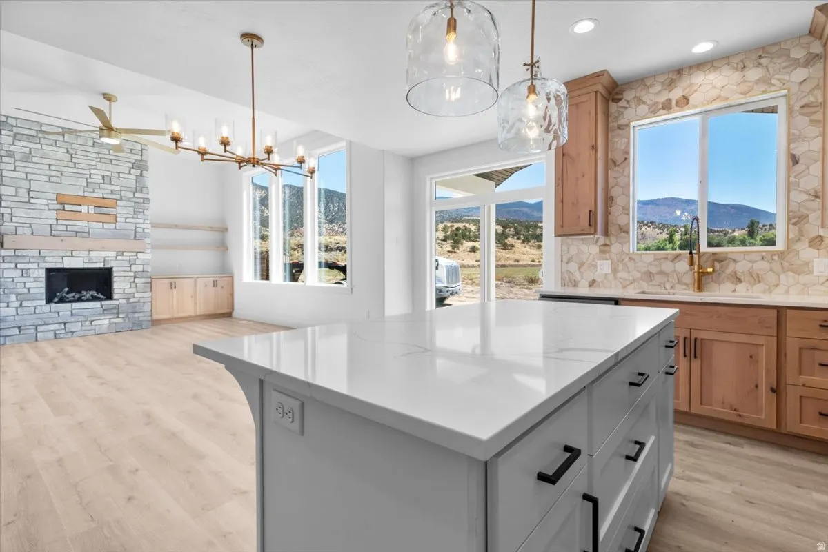 Stone-clad fireplace with wood mantel, large windows, wood-finish flooring, white kitchen island with dark hardware, and a tile backsplash