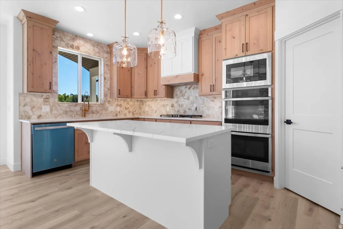 Kitchen featuring a center island with a white countertop, light wood-finish cabinetry, a patterned tile backsplash, stainless steel built-in appliances, and wood-finish flooring