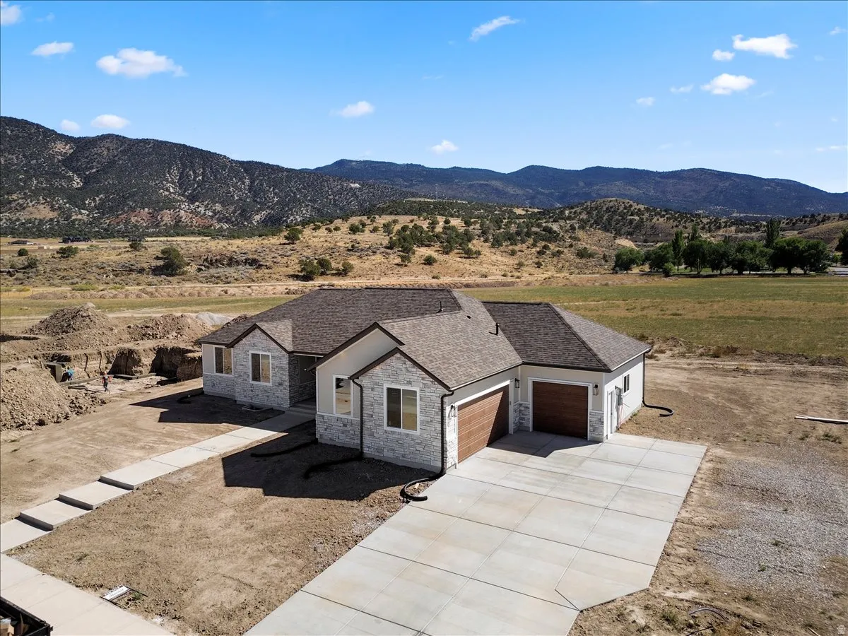 Stone and stucco exterior with a dark-tone shingle roof, wood-finish garage doors, concrete driveway, and mountain backdrop