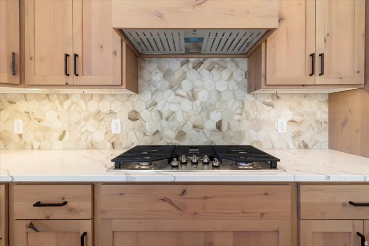 Light wood-finish cabinetry with matte black hardware, built-in cooktop, under-cabinet lighting, and a hexagon tile backsplash