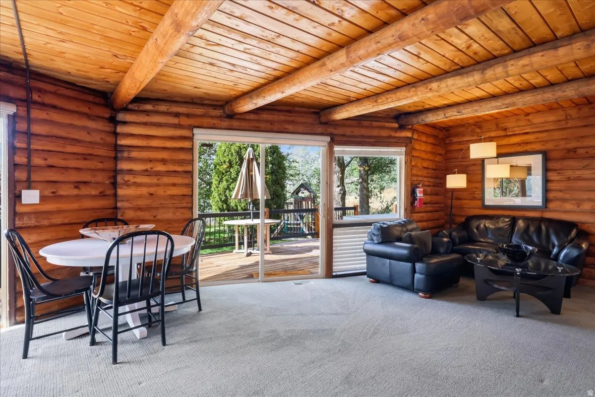 Log-style interior walls with exposed wood ceiling beams, light gray carpet, and a glass sliding door leading to an outdoor deck