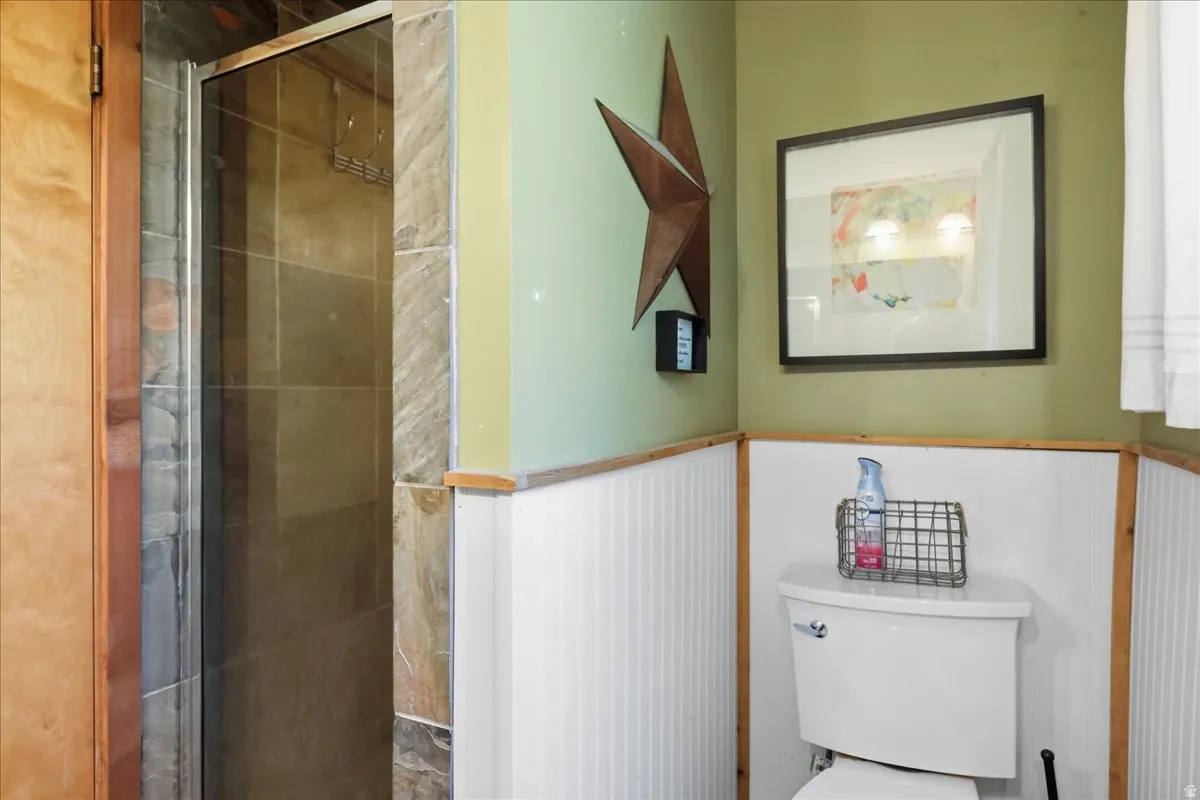 Glass-enclosed shower with tiled surround, white toilet, and wainscoting with wood trim