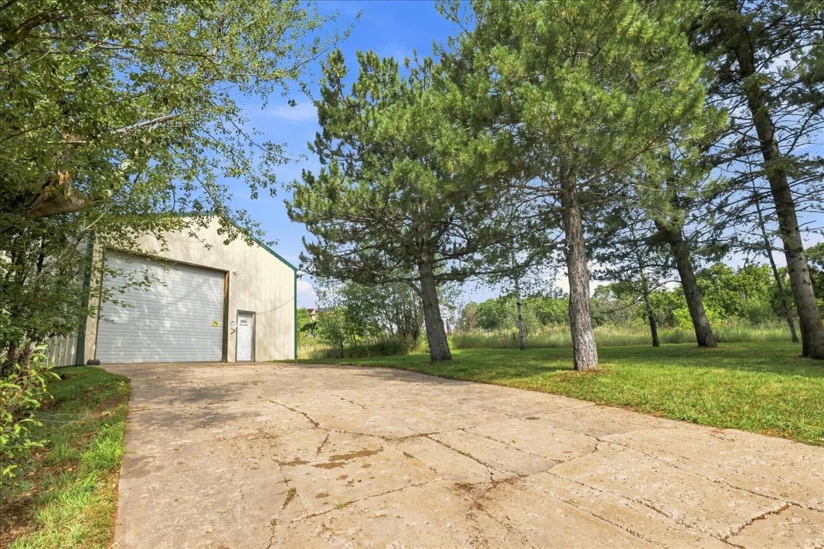 Metal-sided outbuilding with a large roll-up door and separate entry door, a concrete driveway, and mature trees