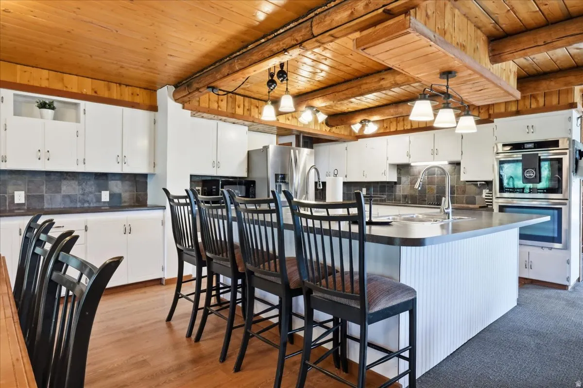 Kitchen featuring a wood-finish ceiling with exposed beams, white cabinetry, dark tile backsplash, a center island with a built-in sink, and stainless steel appliances