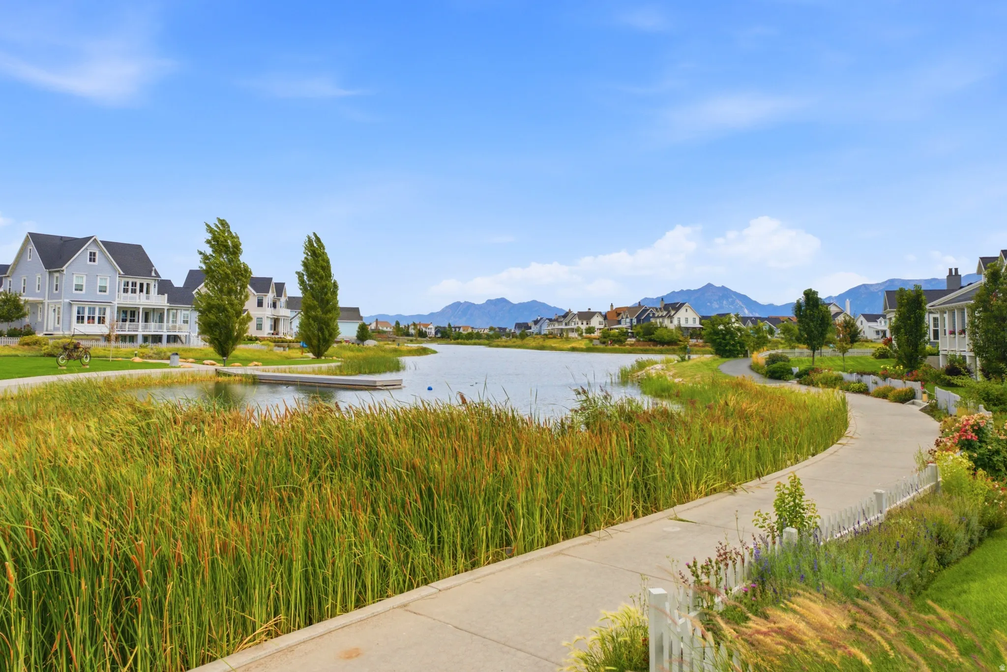 Winding paved pathway bordered by lush greenery and a body of water, with residential architecture in the background