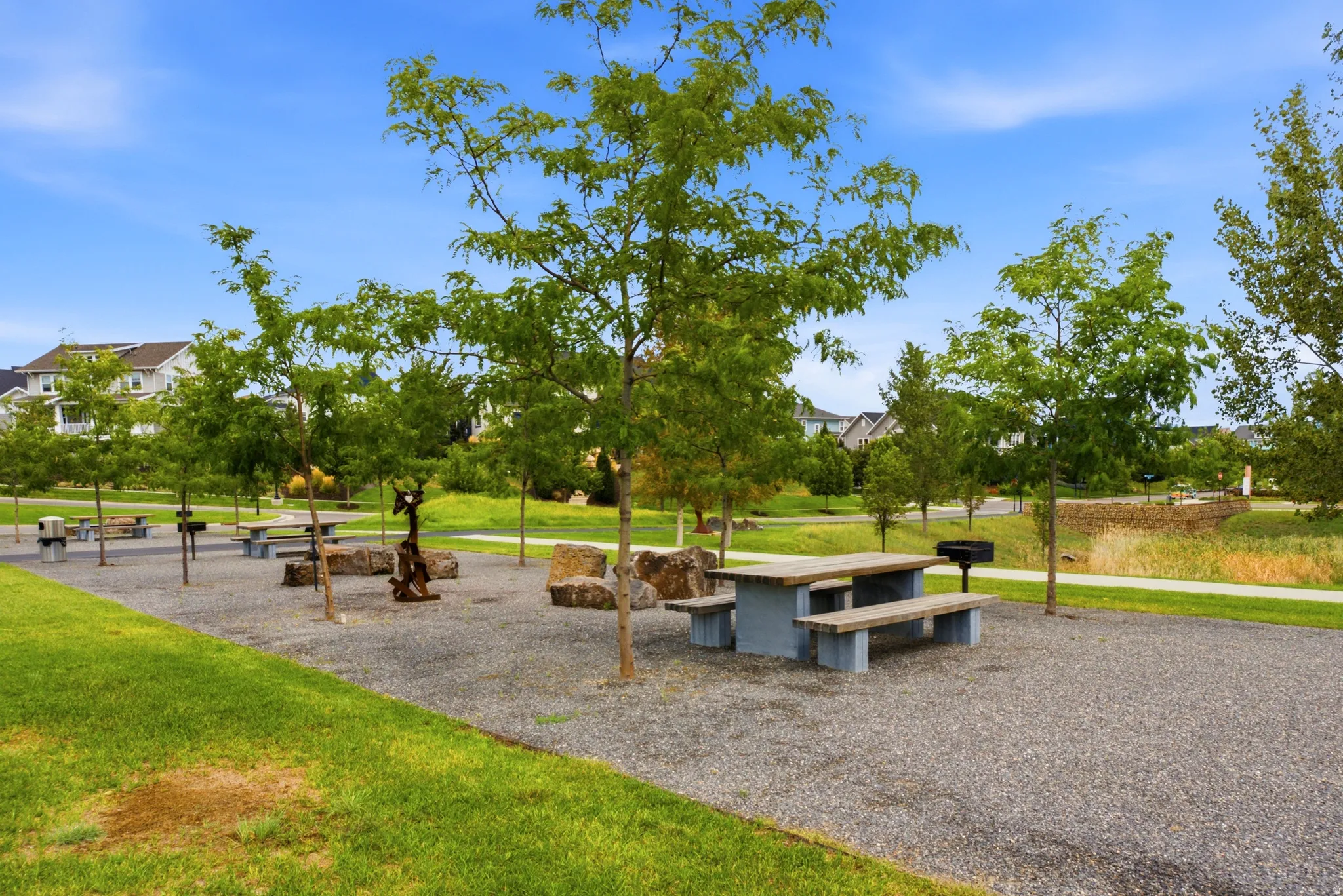 Community park area featuring gravel ground cover, mature trees, and picnic tables with benches