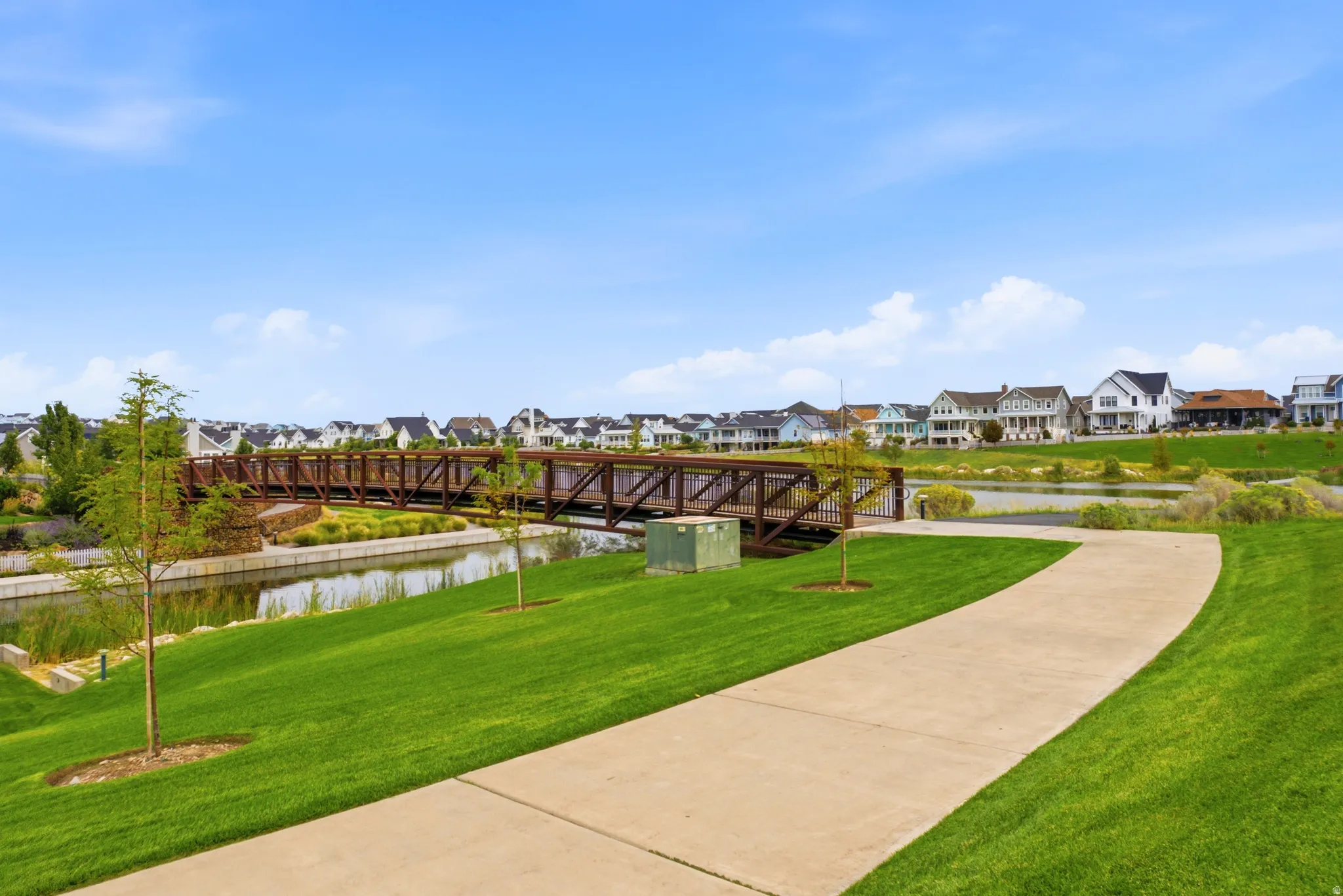 Expansive green space with a paved walking path, mature trees, and a pedestrian bridge over a waterway