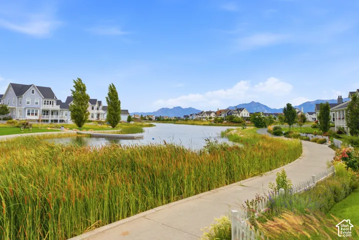 Winding paved pathway bordered by lush greenery and a body of water, with residential architecture in the background