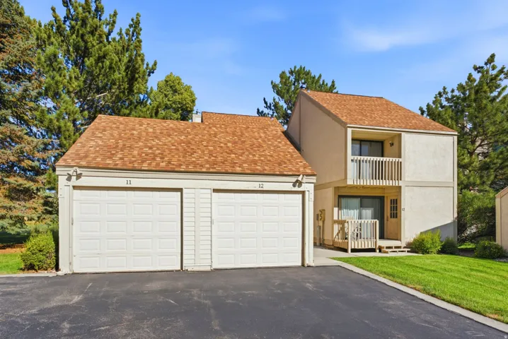Two-car garage with individual doors, asphalt driveway, private upper-level balcony, ground-level patio, and surrounding landscaping