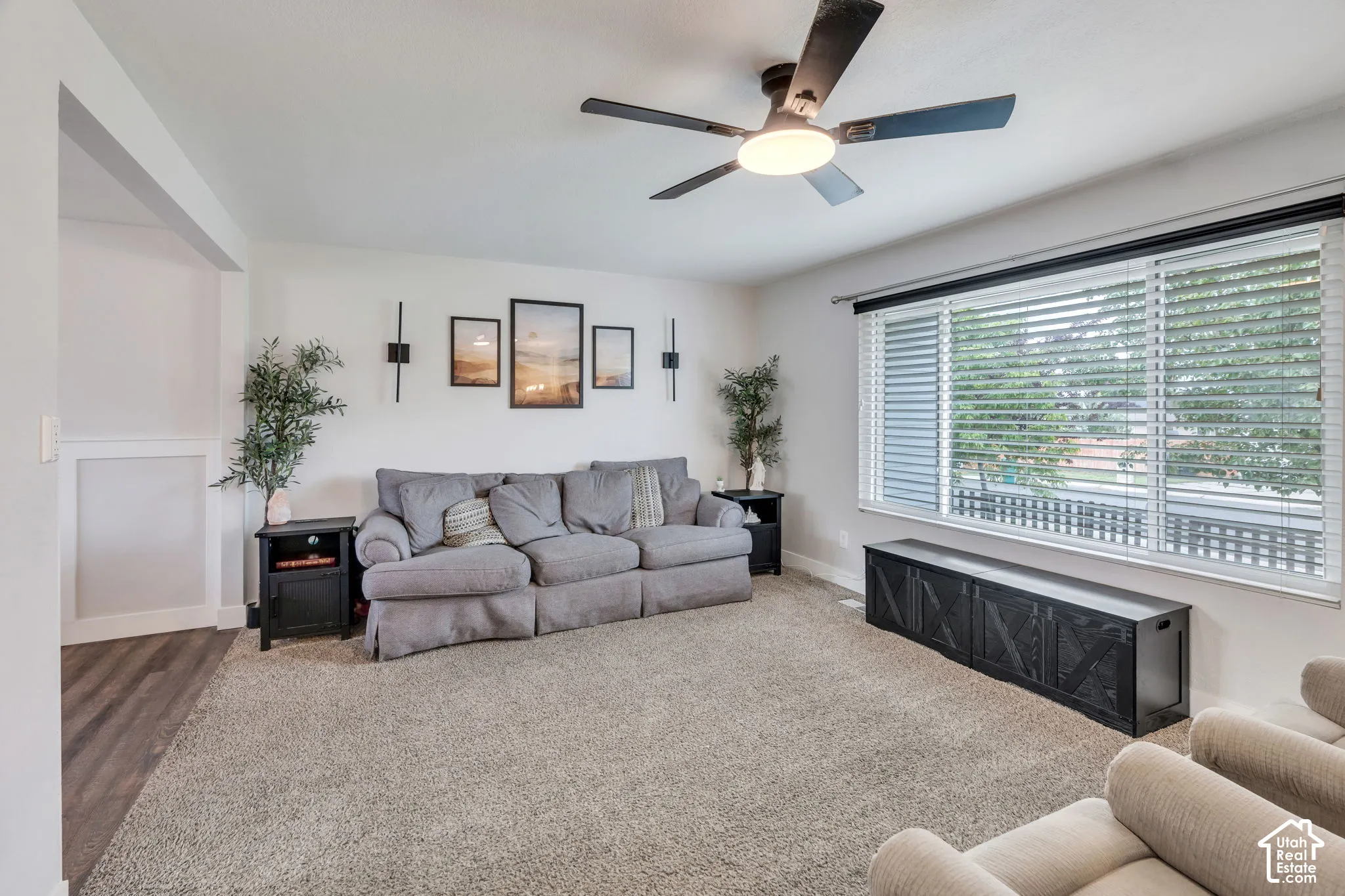 Spacious living area featuring a ceiling fan with integrated lighting, large windows with horizontal blinds, and neutral-toned carpeting