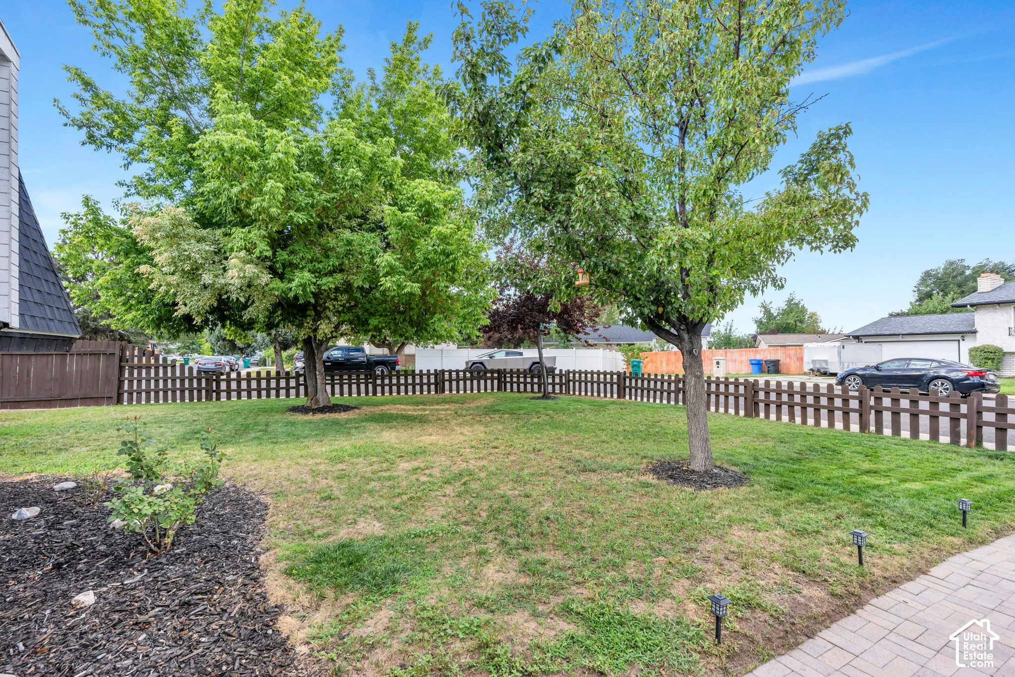 Grassed front yard featuring mature trees, a picket fence, and a paver pathway with integrated lighting