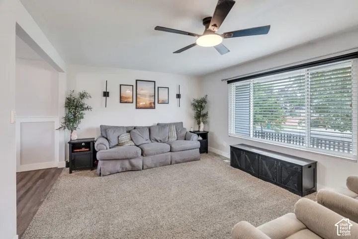 Spacious living area featuring a ceiling fan with integrated lighting, large windows with horizontal blinds, and neutral-toned carpeting
