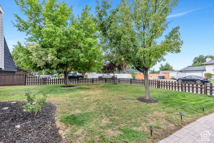 Grassed front yard featuring mature trees, a picket fence, and a paver pathway with integrated lighting