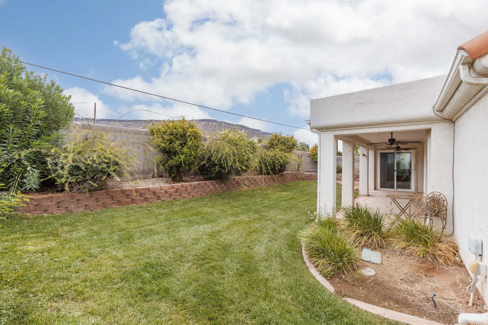 Expansive grass yard with tiered landscaping, mature shrubbery, and a covered patio featuring a ceiling fan